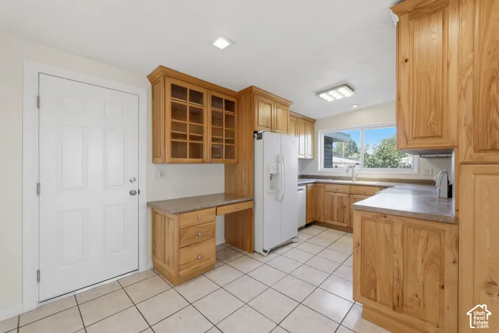 Kitchen featuring natural wood cabinetry, built-in desk area with glass-front upper cabinets, light-toned tile flooring, and integrated white appliances