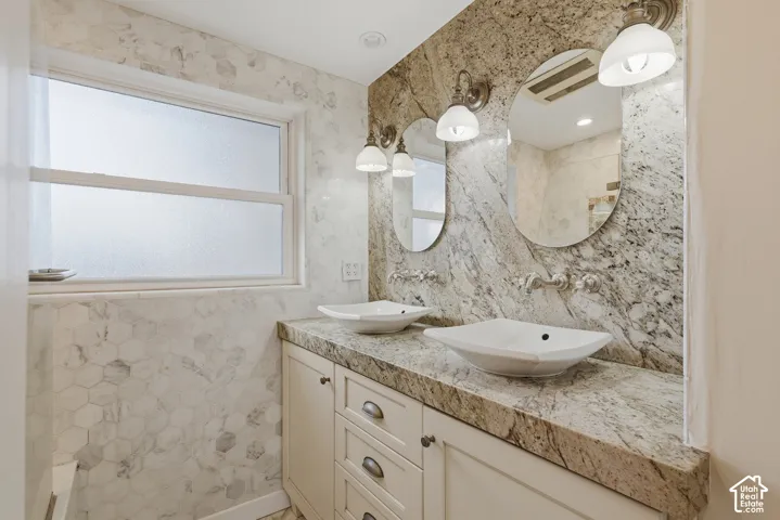 Dual vessel sinks with wall-mounted faucets, stone countertop with matching backsplash, shaker-style cabinetry, and hexagonal wall tile