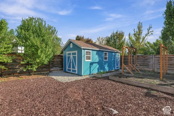 Storage building with dual-entry doors, exterior windows, and shingle roofing, complemented by wood-framed garden beds and enclosed by horizontal wood fencing