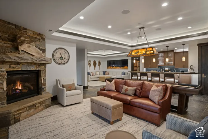Stacked stone fireplace with a thick stone mantel, tray ceiling with recessed lighting, and a built-in wet bar featuring pendant lighting and wood cabinetry