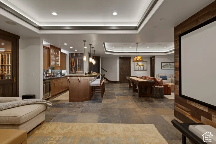Entertainment space featuring stone flooring, a wet bar with cabinetry, custom tray ceilings with recessed lighting, and a wall-mounted projector screen