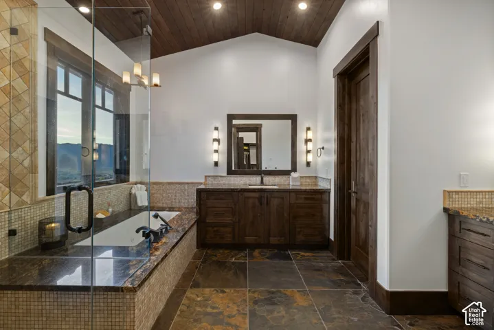 Vaulted wood-plank ceiling, natural stone tile flooring, custom dark wood cabinetry, glass-enclosed shower, and built-in soaking tub with stone surround