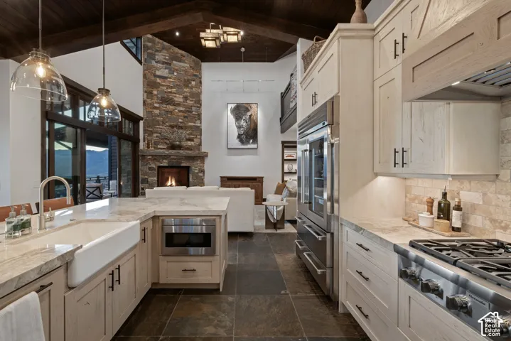 Gourmet kitchen featuring stone countertops, custom cabinetry, a farmhouse sink, stainless steel professional-grade appliances, and slate flooring under a vaulted wood-beam ceiling with a floor-to-ceiling stone fireplace
