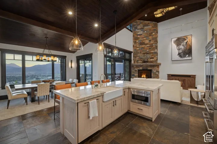 Vaulted timber-plank ceiling, floor-to-ceiling stone fireplace, stone-tile flooring, kitchen island with farmhouse sink, and large-format multi-pane windows