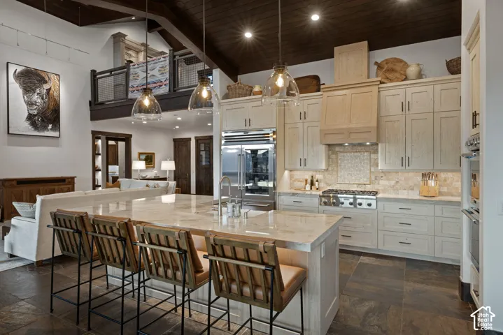 Gourmet kitchen featuring custom light-finish cabinetry, a large central island with stone countertop, stainless steel appliances, a dark wood-plank cathedral ceiling, and stone flooring