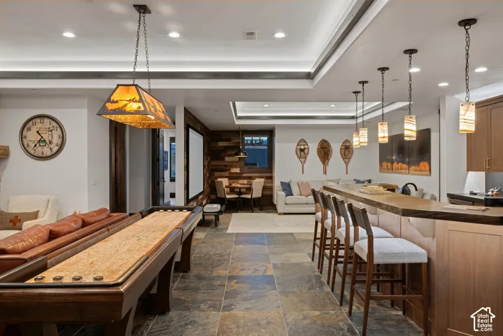 Spacious interior featuring tray ceilings with recessed lighting, large-format stone tile flooring, a custom bar with wood cabinetry, and an accent wall finished in reclaimed wood planks