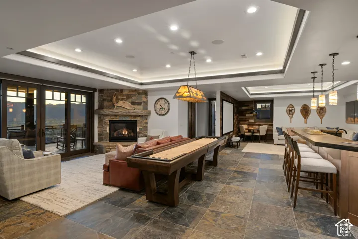 Large living space featuring tile flooring, a tray ceiling with recessed lighting, a stone-clad corner fireplace, and a built-in wet bar with pendant lighting