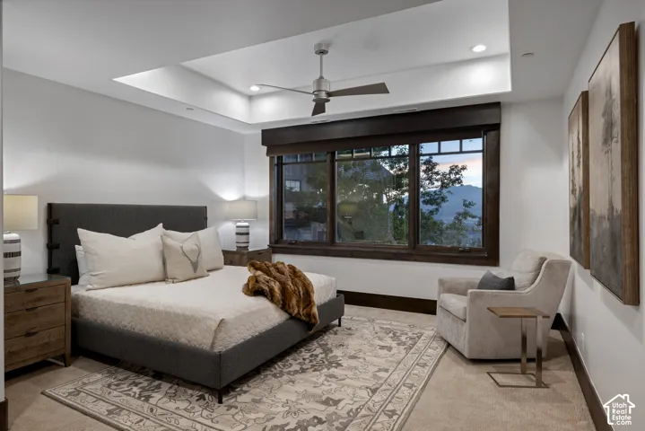 Tray ceiling with a central ceiling fan, large windows with wood-trimmed frames, and carpeted flooring