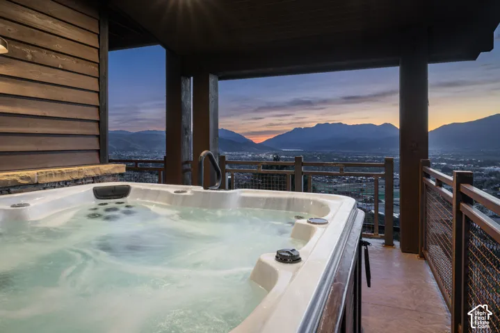 Covered deck featuring a built-in jetted tub, horizontal wood siding, stone base detailing, and metal wire railing overlooking mountain and valley scenery