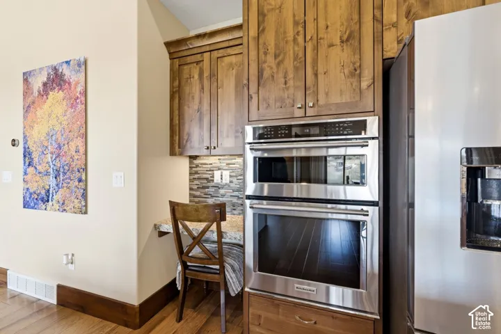 Stained wood cabinetry, stone-tile backsplash, built-in desk area with stone countertop, and stainless steel double wall ovens
