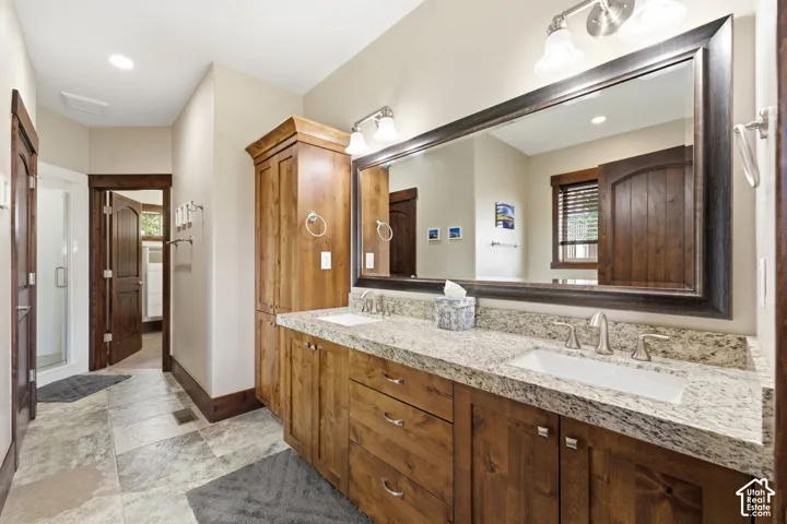 Dual-sink vanity with stone countertop, dark wood cabinetry, tall built-in linen storage, large framed mirror, and tile flooring