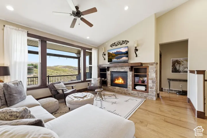 Vaulted ceilings featuring a ceiling fan, stone-surround fireplace with a timber mantel, built-in shelving, wood-finish flooring, and expansive windows