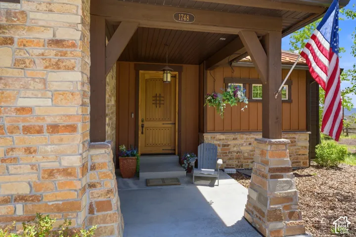 Covered entryway featuring natural stone veneer pillars, vertical wood-panel siding, a solid wood front door with decorative iron hardware, and an integrated ceiling light fixture