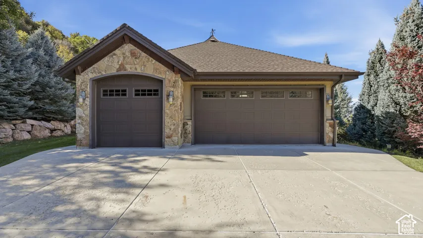 Three-car garage featuring stone masonry exterior accents, dual-bay doors with transom windows, a concrete driveway, and a hipped roofline