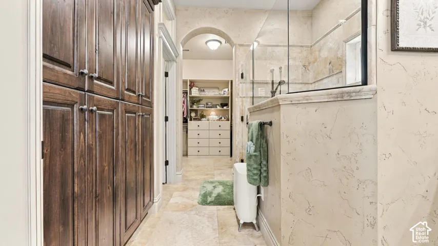 Full-height stained wood cabinetry, natural stone tile flooring, glass-enclosed shower, arched architectural entryway, and built-in closet storage