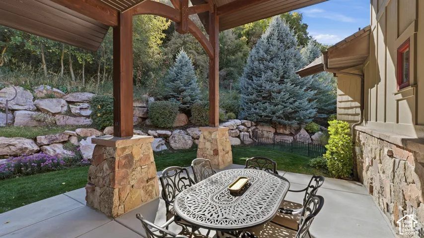 Covered patio featuring timber frame supports, stacked stone column bases, concrete flooring, and a natural stone retaining wall backdrop