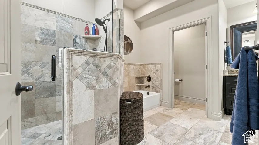 Walk-in shower with stone tile surround, recessed shelving, and a glass partition, complemented by stone tile flooring, a built-in bathtub, and a dark-finish vanity with a stone countertop