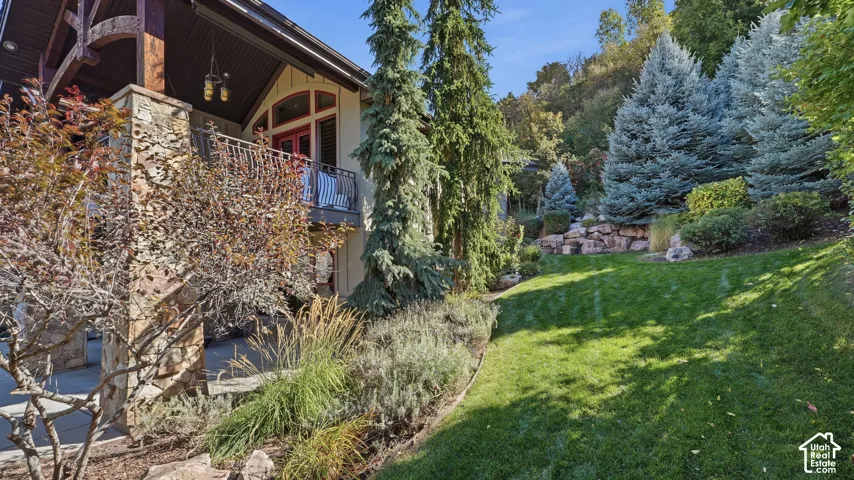 Stone masonry exterior detailing, covered patio, wrought iron balcony railing, manicured lawn, and tiered rock landscaping