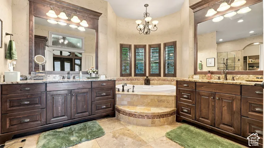 Dual wood-finish vanities with stone countertops and framed mirrors, central jetted soaking tub with stone tile surround, arched window array, and overhead chandelier lighting