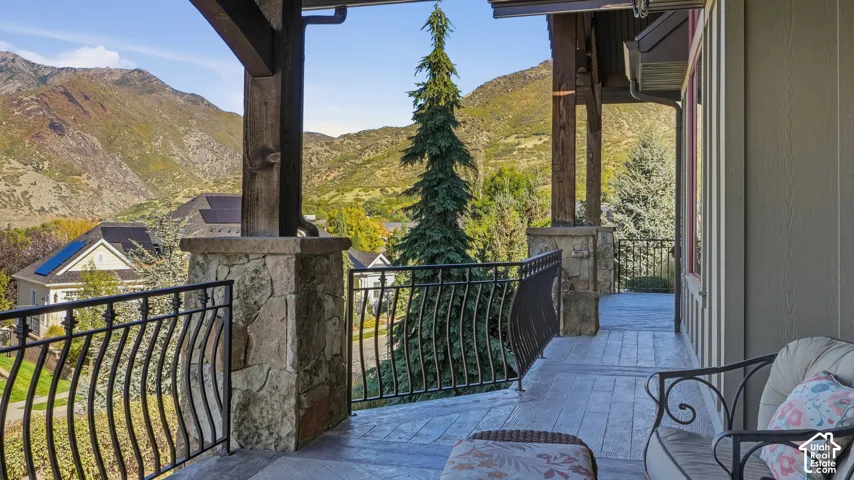 Covered porch featuring wood-grain stamped concrete flooring, exposed timber framing, stone-clad pillar bases, and decorative metal railings with mountain scenery backdrop