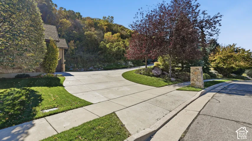 Concrete driveway featuring a stone-clad pillar, manicured lawn, and mature landscaping against a hillside backdrop