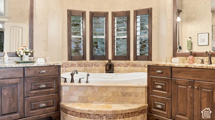 Built-in soaking tub with a stone-tiled surround, dual stained-wood vanities featuring stone countertops, and four windows with wood shutters