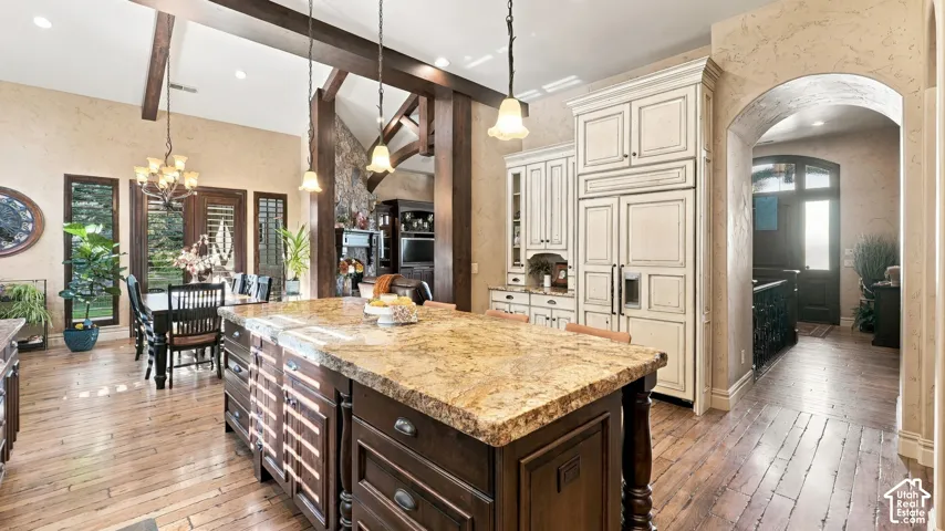 Kitchen featuring a stone-topped island with base cabinetry, custom panel-ready refrigerator, wood-finish flooring, vaulted ceilings with exposed structural beams, and a stone-accented fireplace