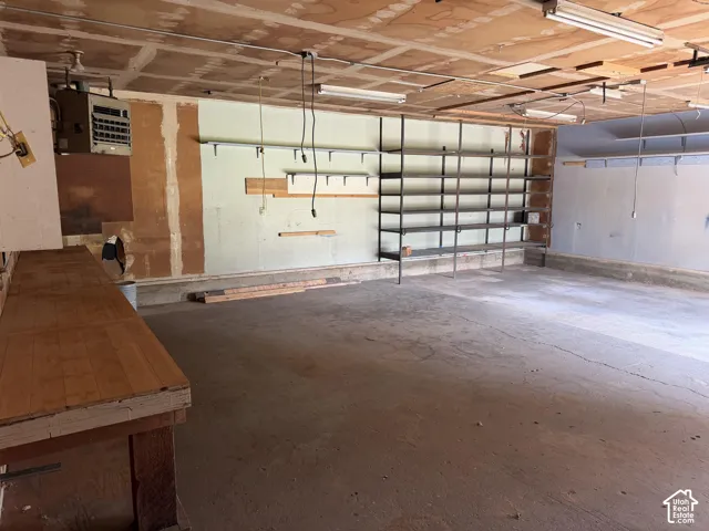 Garage space featuring a concrete floor, built-in wooden workbench, metal industrial shelving, exposed ceiling lighting, and wall-mounted heater unit