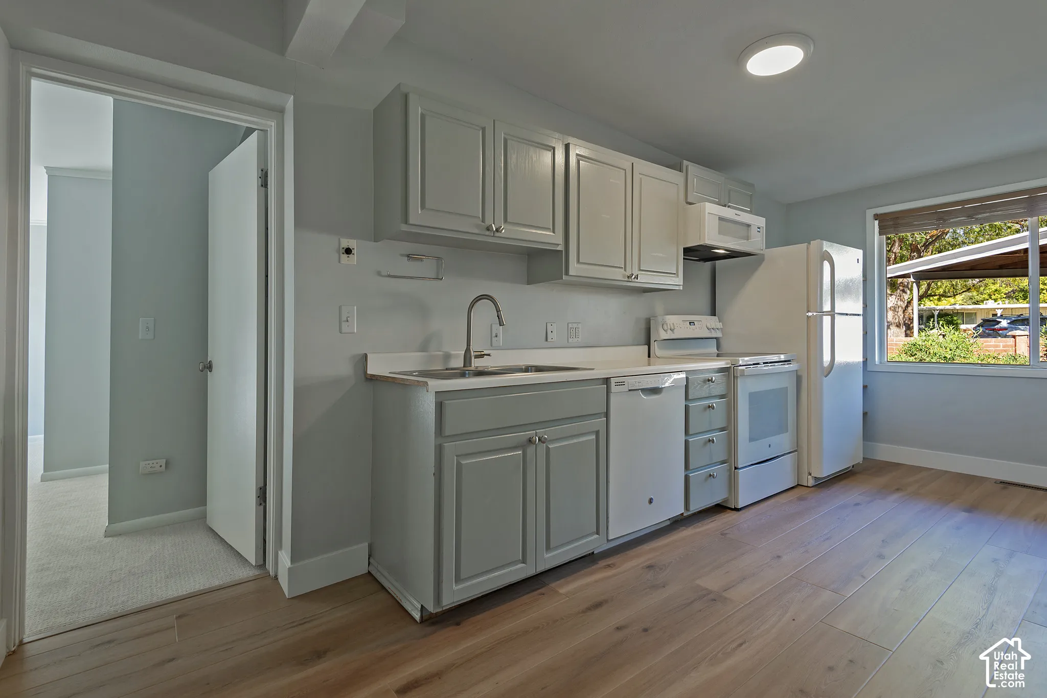 Kitchen featuring wood-finish flooring, shaker-style cabinetry, a stainless steel dual-basin sink with a gooseneck faucet, and white appliances including a built-in dishwasher and microwave