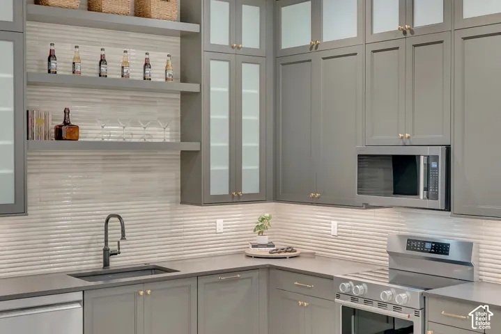 Custom cabinetry with glass-panel upper doors, integrated stainless steel appliances, floating wood-finish shelving, textured tile backsplash, and a matte black gooseneck faucet