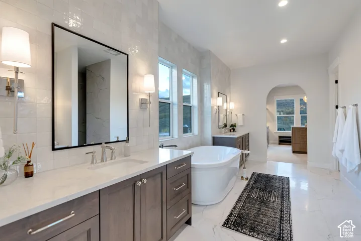 Double vanity featuring wood-finish cabinetry, quartz countertops, and metallic wall-mounted sconces, paired with a freestanding soaking tub, tiled wall accents, and stone-finish flooring