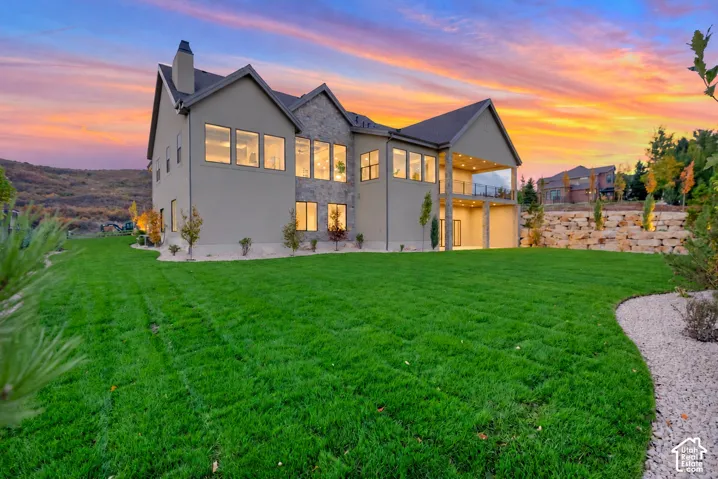 Exterior rear facade featuring stone masonry accents, a multi-level covered patio with glass railings, numerous large windows, and a landscaped backyard with decorative stone retaining walls