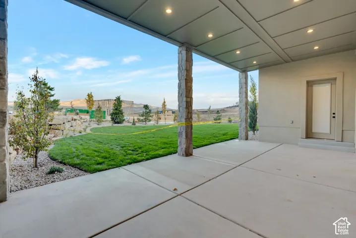 Covered concrete patio featuring recessed ceiling lighting and stone-clad support columns, opening onto a landscaped backyard with rock wall elements
