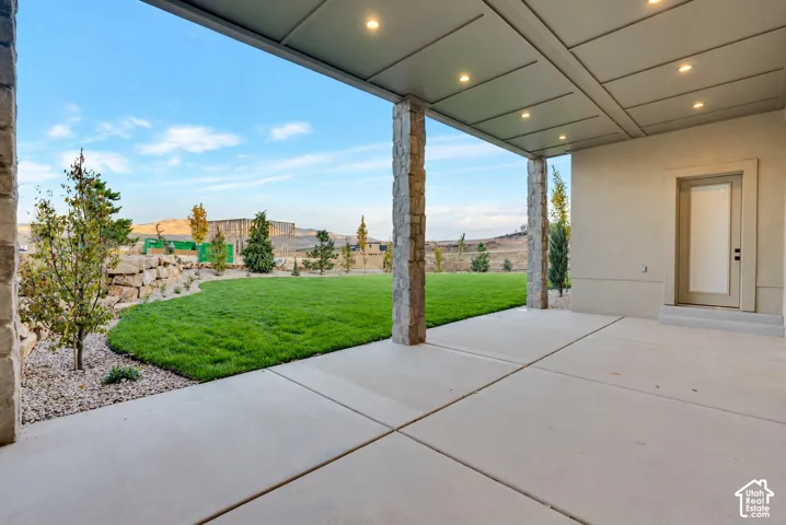 Covered concrete patio featuring recessed lighting and natural stone columns, overlooking a manicured lawn with decorative rock landscaping and retaining walls
