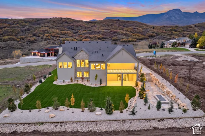 Multi-story residence featuring stone and stucco exterior, covered balcony with glass railing, manicured lawn, flagstone pathway, and stone-retaining walls set against a mountain backdrop