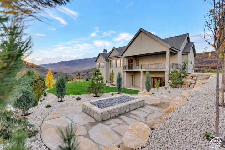Exterior featuring a stone-clad fire pit, multi-level flagstone patio, covered deck with iron railing, manicured lawn, and mountainous horizon