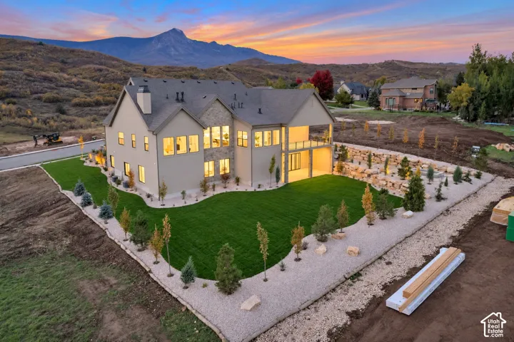 Multi-story exterior featuring stone and stucco siding, a covered upper-level deck, expansive windows, manicured lawn, and mountain backdrop