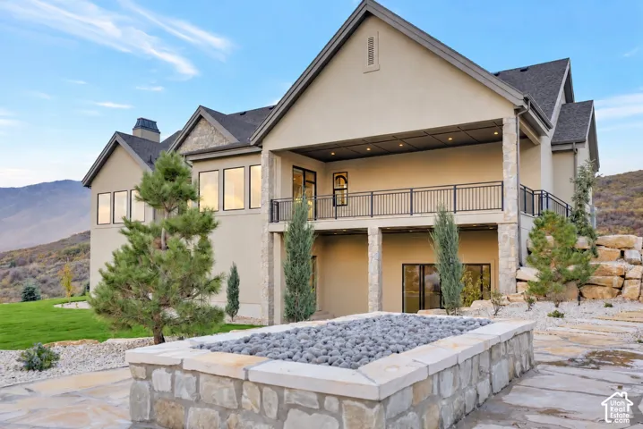 Two-story exterior featuring stone veneer accents, an expansive covered balcony with black metal railings, a stone-surround fire feature, and natural rock retaining walls