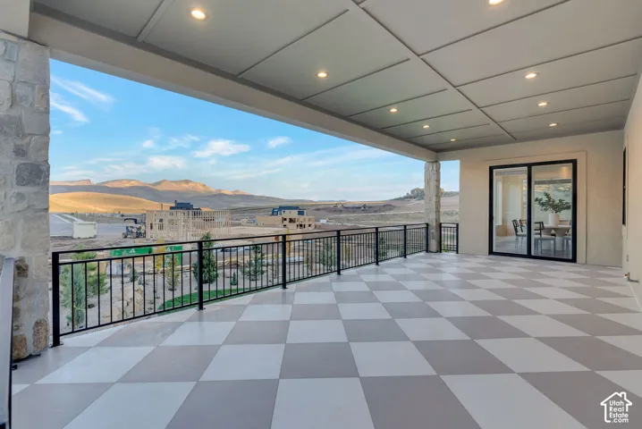 Covered patio featuring a checkered tile floor, recessed lighting, stone-clad pillars, and a black metal railing overlooking mountainous terrain
