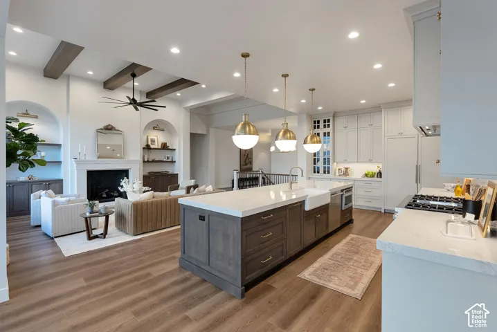 Kitchen featuring a large central island with quartz countertops, custom cabinetry, apron-front sink, and pendant lighting, complemented by wood-finish flooring, exposed ceiling beams, and a fireplace with arched built-in shelving