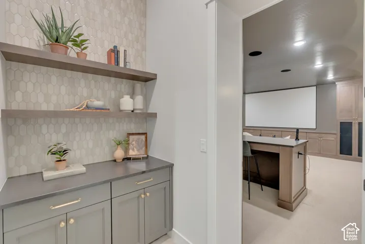 Custom wet bar featuring matte gray cabinetry, brass hardware, stone-finish countertops, floating wood shelves, and a picket-pattern tile backsplash