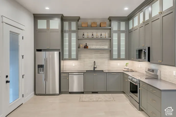 Custom cabinetry with glass-front uppers, stainless steel appliances, open floating shelving, horizontal tile backsplash, and dark-toned hardware