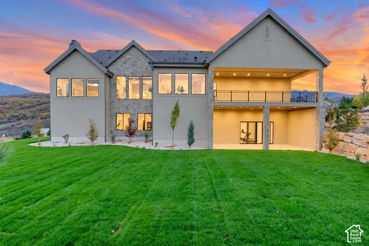 Multi-story exterior featuring stone veneer accents, an expansive rear lawn, a covered walk-out patio, and an elevated balcony with metal railings