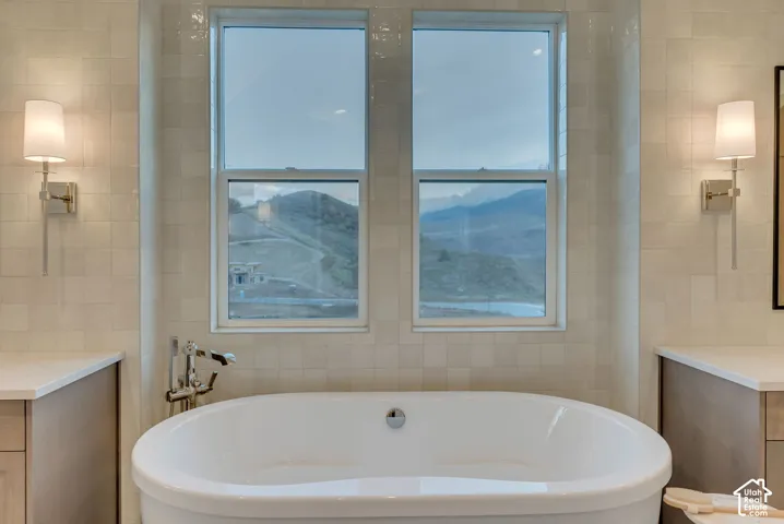 Freestanding white soaking tub with polished chrome wall-mounted faucet, offset by dual polished chrome wall sconces and a mountain backdrop