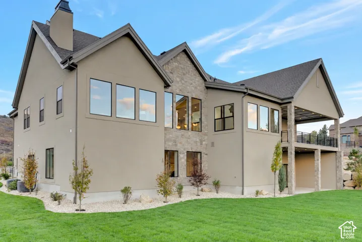 Multi-level residence featuring stucco siding, stone veneer accents, large picture windows, a covered upper-level deck with metal railing, and an expansive lawn