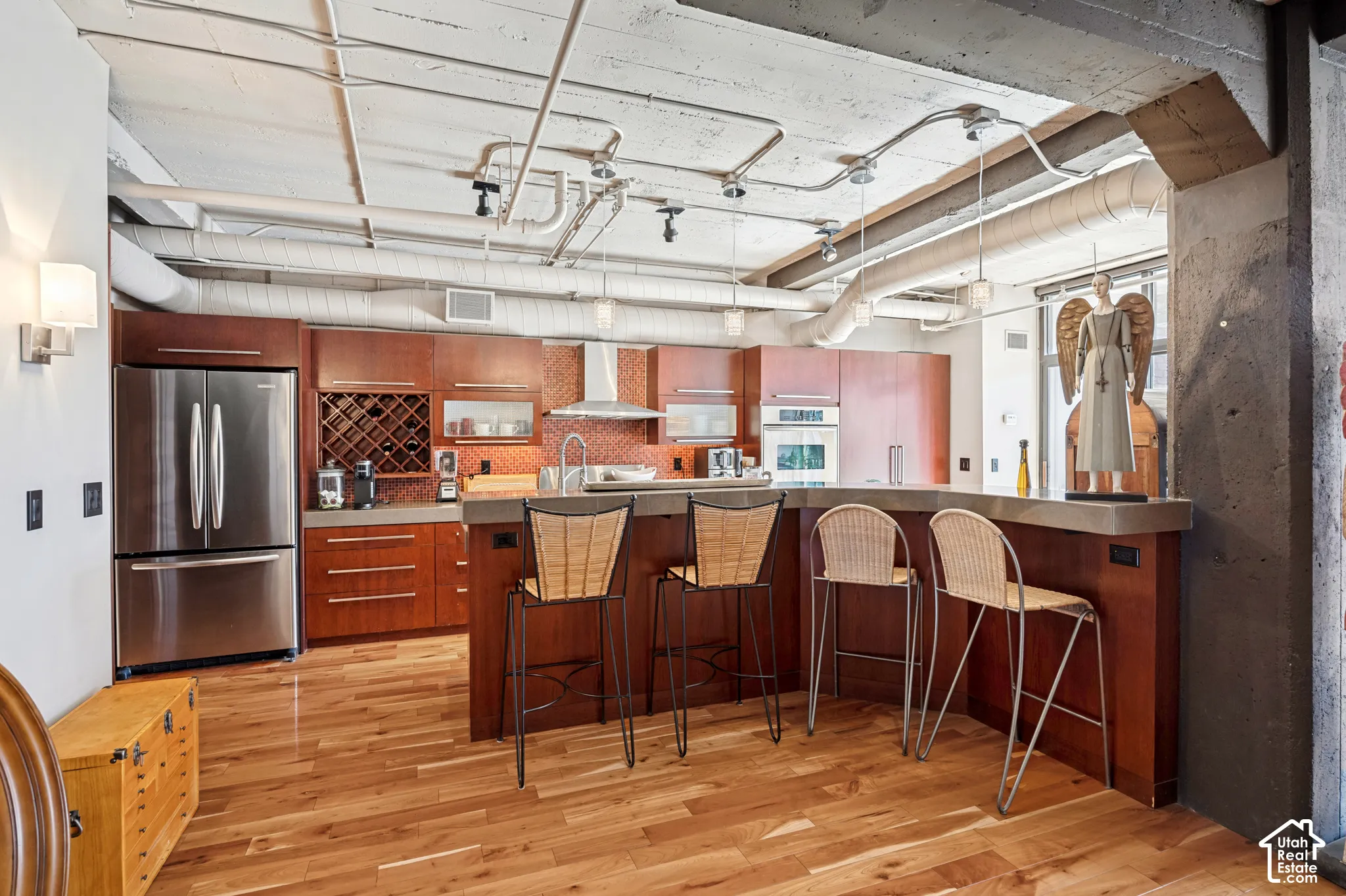 Industrial-style kitchen featuring exposed concrete ceilings and utility piping, custom wood-finish cabinetry, solid-surface breakfast bar, mosaic tile backsplash, and wood-finish flooring