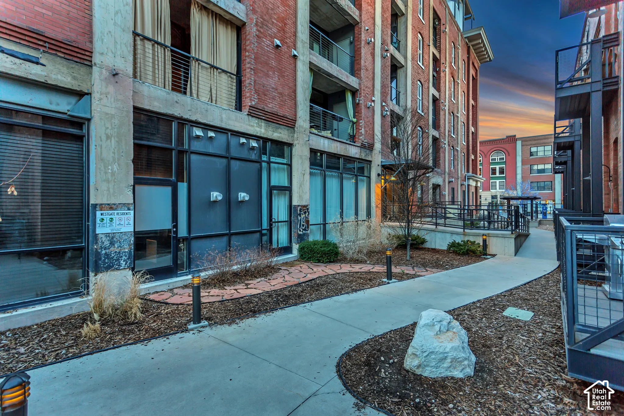 Industrial-style building exterior featuring exposed brick facade, expansive metal-framed window systems, concrete columns, integrated balconies, and a paved walkway with stone hardscaping