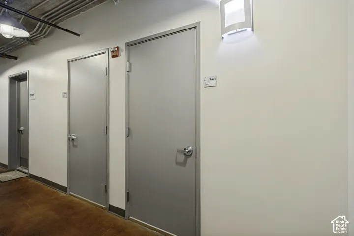 Industrial-style hallway featuring concrete flooring, exposed overhead piping, minimalist slab doors, and architectural wall-mounted sconces