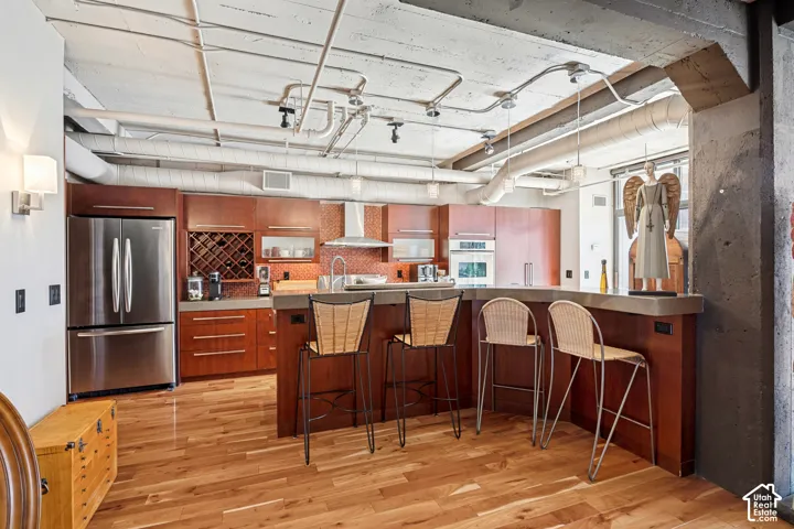 Industrial-style kitchen featuring exposed concrete ceilings and utility piping, custom wood-finish cabinetry, solid-surface breakfast bar, mosaic tile backsplash, and wood-finish flooring