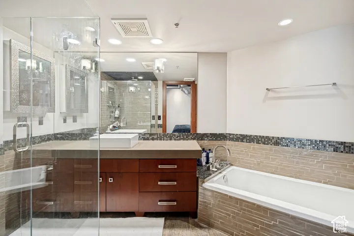 Bathroom featuring a custom wood-finish vanity with a rectangular vessel sink, expansive wall-to-wall mirror, glass-enclosed shower, and a built-in soaking tub surrounded by linear stone tile and mosaic accents